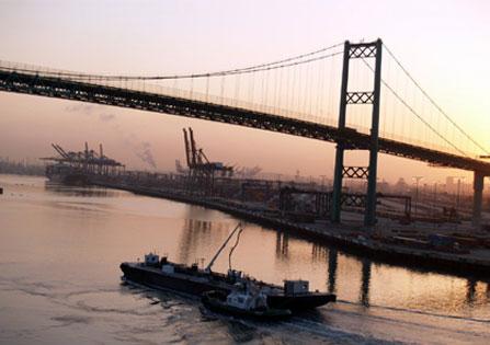(Photo by wirralwater @ Flickr) Cranes silhouetted in an industrial smog mist.  In the foreground a tug escorts a barge beneath a large suspension bridge at the Port of Los Angeles.